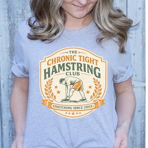 May include: A light gray t-shirt featuring a graphic for "The Chronic Tight Hamstring Club." The design includes a person stretching, laurel leaves, stars, and the text "Stretching Since 2003."
