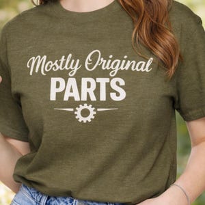 May include: An olive green t-shirt featuring the white text "Mostly Original PARTS". A gear graphic is centered below the text. The shirt is made of a soft, comfortable material.