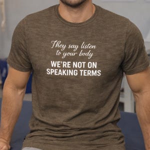 Op de afbeelding: Bruin t-shirt met de witte tekst: "They say listen to your body, we're not on speaking terms". Het t-shirt wordt door een persoon gedragen. Het t-shirt heeft een gemêleerde bruine kleur.