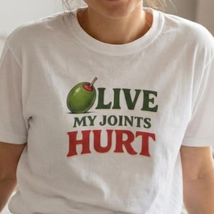 Puede incluir: Camiseta blanca con un gráfico de aceituna verde y el texto "LIVE MY JOINTS HURT" en verde y rojo. La camiseta tiene cuello redondo y mangas cortas.