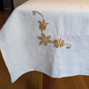 May include: Off-white rectangular tablecloth with embroidered floral design in shades of gold. The tablecloth is draped over a wooden table, showcasing the corner with the embroidered flowers and leaves. The edge of the tablecloth has a gold trim.