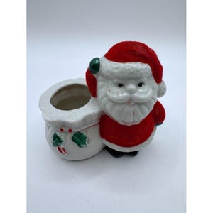 May include: A ceramic Santa Claus figurine featuring a red flocked hat and suit, standing next to a white sack with a hollow opening. The sack is decorated with painted green holly leaves and red berries on the front, with a smooth white finish.