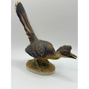 Peut inclure: Une figurine de géocoucou en céramique avec une longue queue dressée, une crête texturée sur la tête et un bec pointu. L'oiseau est peint dans des tons de brun, gris et beige avec une petite tache orange près de l'œil, posé sur un socle.