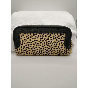 Puede incluir: Cartera rectangular con marco de cuero negro y centro moteado marrón y negro. La cartera tiene una cremallera dorada y un borde de cuero negro. La cartera está sobre una superficie blanca.