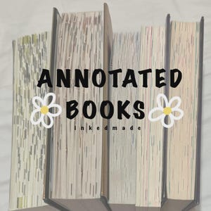 May include: A stack of books with the words "ANNOTATED BOOKS" in black text, with a white and yellow flower graphic on either side. The text "inkedmade" is below the word "BOOKS". The books have light-colored pages.
