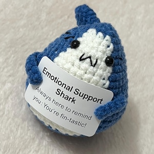 May include: A handmade blue and white crocheted shark holding a sign. The sign says "Emotional Support Shark" and "Always here to remind you: You're fin-tastic!" The shark has black eyes and a white belly.