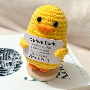 May include: A handmade yellow crochet duck figurine holding a sign that reads "Positive Duck". The duck has black eyes and an orange beak and feet. The sign says "I may be a tiny duck, but I believe in you! Go do your thing! You're Ducking Awesome!"
