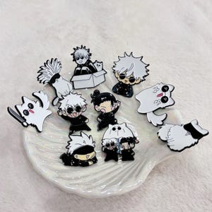 May include: A collection of enamel pins featuring anime-style characters in black and white. The pins depict various characters with spiky white hair, sunglasses, and cat-like figures. The pins are arranged on a white, shell-shaped dish.