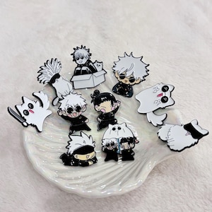 Op de afbeelding: Een verzameling emaille pins met anime-stijl karakters in zwart en wit. De pins tonen verschillende karakters met stekelig wit haar, katten en andere accessoires. De pins zijn gerangschikt op een witte, schelpvormige schaal.