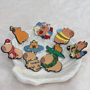 Puede incluir: Una colección de pines de esmalte con capibaras en varios diseños. Un pin muestra un capibara con gorro y bufanda de Papá Noel, mientras que otro está en un scooter. Otros pines incluyen un capibara en una piscina y uno con una corona navideña.