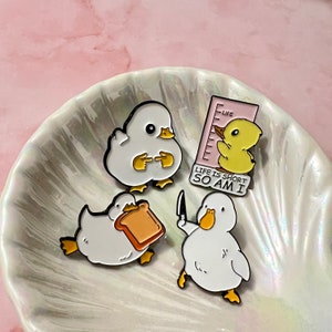 Könnte beinhalten: Vier Emaille-Pins mit Cartoon-Enten. Eine Nadel ist eine gelbe Ente mit dem Text "Life is short so am I". Eine andere Nadel ist eine weiße Ente, die ein Stück Brot hält. Die beiden anderen Nadeln sind weiße Enten mit verschiedenen Posen.