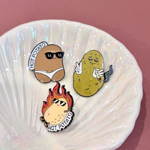 Puede incluir: Tres broches de esmalte novedosos con patatas antropomórficas. Uno es marrón con gafas de sol y braguita blanca, otro es amarillo con un signo de la paz y un cigarrillo encendido, y el tercero es una patata en llamas con gafas de sol. Cada pin tiene la inscripción "HOT POTATO".