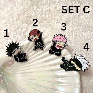 May include: Set of four black and white anime character enamel pins. The pins are on a white seashell-shaped surface. The pins are numbered 1 through 4.