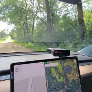 May include: A view from inside a car driving down a dirt road through a forest. The car's dashboard is visible, with a laptop displaying a map and a digital clock showing 20. The road is lined with trees on both sides.