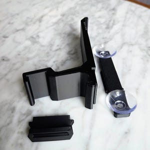 May include: Black plastic car phone mount with a suction cup and adjustable arm. The mount has a cradle to hold the phone and a base that attaches to the dashboard. The mount is designed to hold a phone securely while driving.