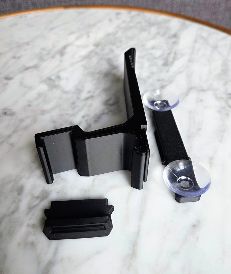 May include: Black plastic car phone mount with a suction cup and adjustable arm. The mount has a cradle to hold the phone and a base that attaches to the dashboard. The mount is designed to hold a phone securely while driving.