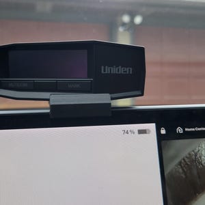 May include: A black Uniden radar detector mounted above a vehicle's digital dashboard screen. The device features a dark display screen and two buttons labeled MUTE/DIM and MARK. The dashboard screen displays vehicle status icons, time, and temperature.