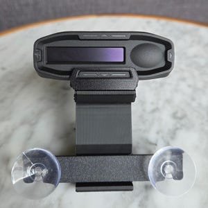 May include: A black electronic device with a rectangular screen and a control button. The device is mounted on a bracket with two clear suction cups. The device appears to be a car accessory.