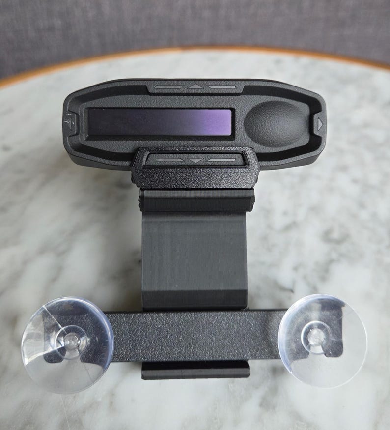May include: A black electronic device with a rectangular screen and a control button. The device is mounted on a bracket with two clear suction cups. The device appears to be a car accessory.