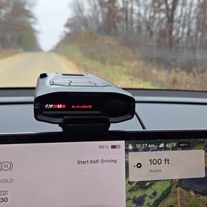 May include: A black radar detector is mounted on a car dashboard, displaying "1.9 U AutoMnv" in red. The car's screen shows "Start Self-Driving" and a map with a distance of 30.48 metres. The car is on a dirt road.