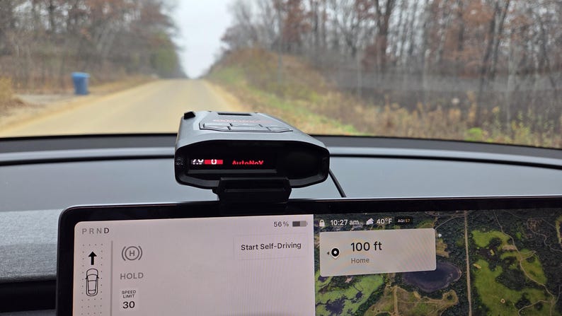 May include: A black radar detector is mounted on a car dashboard, displaying "1.9 U AutoMnv" in red. The car's screen shows "Start Self-Driving" and a map with a distance of 30.48 metres. The car is on a dirt road.