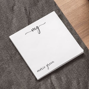 May include: A white notepad with the initials "mg" written in cursive and the text "marie green" printed below. The notepad is on a grey surface.