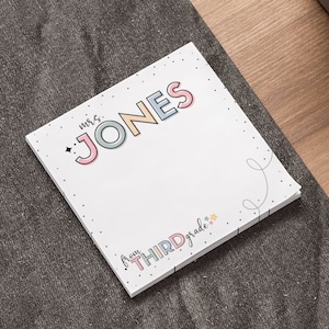 May include: A white note pad with the text "Mrs. JONES" in colorful letters and "from THIRD grade" in smaller colorful letters. The note pad is on a gray surface.