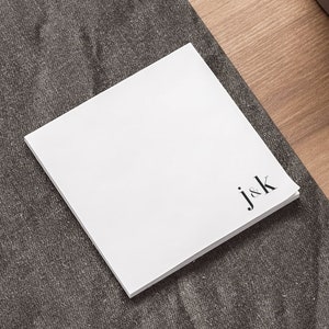 May include: A stack of white square sticky notes with the initials "j&k" printed in black in the lower right corner. The notes are on a dark gray desk mat, with a laptop and pen visible in the background.