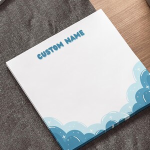 Puede incluir: Un bloc de notas blanco con un diseño de nubes azules y el texto "CUSTOM NAME" en azul. El bloc de notas está sobre una superficie de madera junto a un portátil y un bolígrafo.
