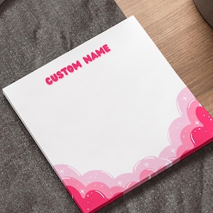 May include: A white notepad with a pink cloud design along the bottom edge. The words "CUSTOM NAME" are printed in pink at the top. The notepad is on a wooden surface next to a laptop and a pen.
