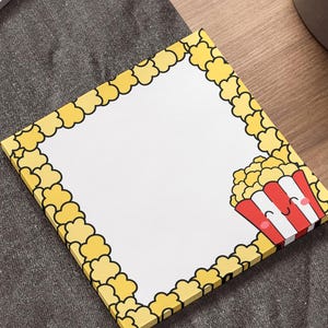 May include: Square notepad with a popcorn theme. The border is yellow popcorn kernels, and a cartoon popcorn container is in the corner. The notepad is on a wooden surface next to a laptop and a plant.