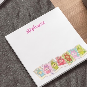May include: A white notepad with the name "Stephanie" in pink script. The bottom edge features a series of colourful cartoon character illustrations in a polaroid style. The notepad is on a dark grey surface, with a laptop and pen visible.