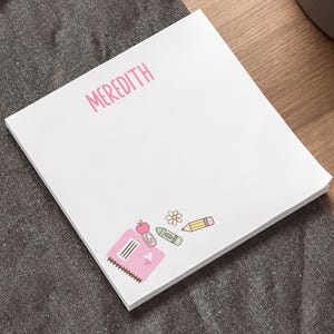 May include: A white square notepad with the name "MEREDITH" in pink script. The notepad features a pink school-themed graphic with a pencil, flower, and apple. The notepad is on a wooden desk next to a laptop and a pen.