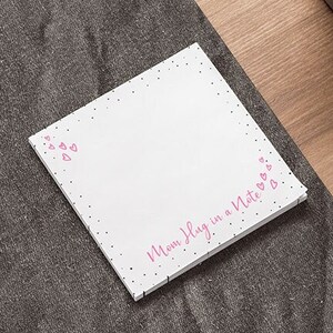 May include: A white notepad with pink hearts and the text "Mom Hug in a Note" printed on it. The notepad is on a grey surface with a laptop and a potted plant in the background.