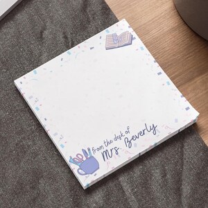 May include: A white notepad with a pink, blue, and white confetti border. The notepad has the text "from the desk of Mrs. Beverly" printed on it. There is a drawing of a mug with pencils and a book on the notepad.