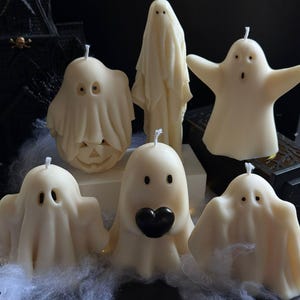 May include: A collection of ivory-colored ghost-shaped candles with various expressions. Some have simple eye cutouts, while others feature open mouths or a heart-shaped detail. The candles are arranged on a dark surface with decorative cobwebs, evoking a Halloween theme.
