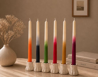 Könnte beinhalten: Ein Set aus sechs hohen Stabkerzen, die jeweils eine weiße Spitze haben, welche in eine einfarbige Basis in Orange, Dunkelblau, Grün, Rot, Senfgelb oder Magenta übergeht. Jede Kerze steht aufrecht in einem strukturierten, cremeweißen Kerzenhalter.