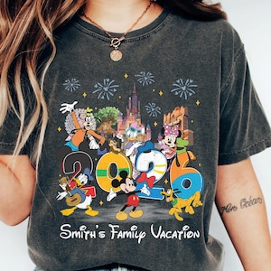 Może przedstawiać: Ciemnoszara koszulka z kolorową grafiką postaci Disneya, w tym Myszki Miki, Kaczora Donalda i Myszki Minnie, z rokiem "2026" i napisem "Smith's Family Vacation". Projekt zawiera fajerwerki i zamek.
