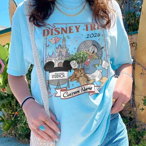Pu&ograve; includere: T-shirt azzurra con un design a tema viaggio Disney con la scritta "Disney Trip 2026". La grafica include Paperino, il castello di Cenerentola e il logo Hollywood Studios. Un banner recita "Custom Name".