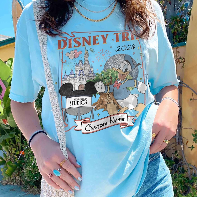 Pu&ograve; includere: T-shirt azzurra con un design a tema viaggio Disney con la scritta "Disney Trip 2026". La grafica include Paperino, il castello di Cenerentola e il logo Hollywood Studios. Un banner recita "Custom Name".