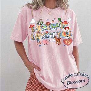 May include: A light pink short-sleeve t-shirt featuring a colorful graphic print of Toy Story characters including Woody, Buzz Lightyear, Jessie, Rex, and Bullseye. The text reads I KNEW IT I KNEW YOU with a red heart containing the letters TS.