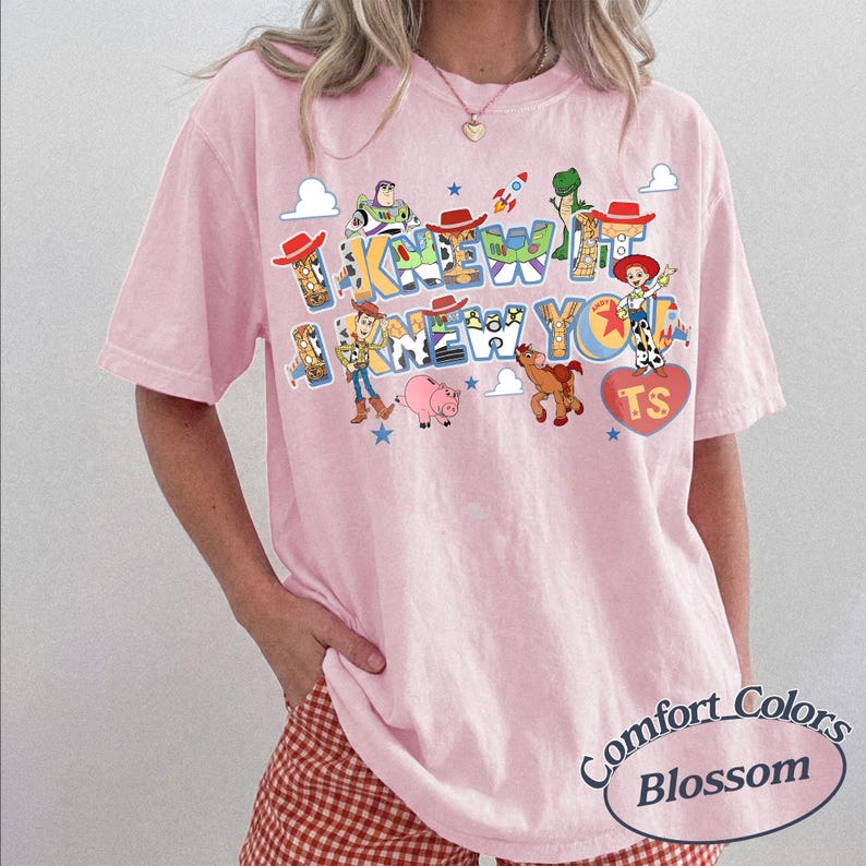 May include: A light pink short-sleeve t-shirt featuring a colorful graphic print of Toy Story characters including Woody, Buzz Lightyear, Jessie, Rex, and Bullseye. The text reads I KNEW IT I KNEW YOU with a red heart containing the letters TS.