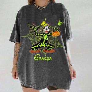 May include: Dark gray t-shirt featuring a cartoon character in a skeleton costume holding a jack-o'-lantern. The character is set against a green spiderweb design with the word "Grandpa" in green script at the bottom. A green spider is in the upper right corner.