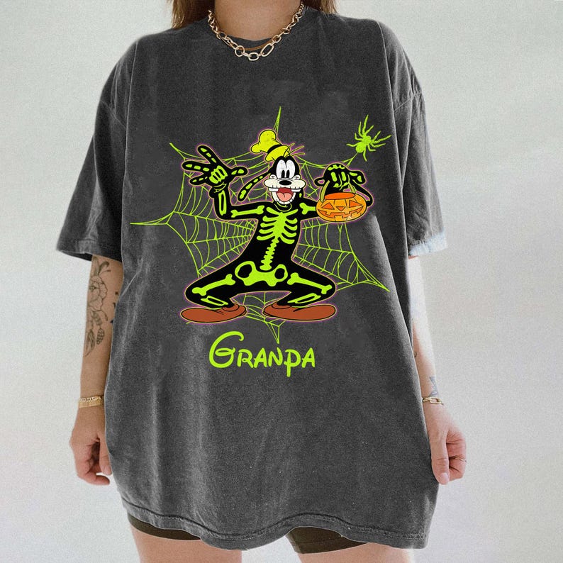 May include: Dark gray t-shirt featuring a cartoon character in a skeleton costume holding a jack-o'-lantern. The character is set against a green spiderweb design with the word "Grandpa" in green script at the bottom. A green spider is in the upper right corner.