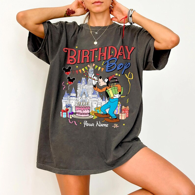 K&ouml;nnte beinhalten: Dunkelgraues T-Shirt mit einer farbenfrohen Grafik, die eine Zeichentrickfigur, ein Schloss und die Worte "Birthday Bop" zeigt. Das Design umfasst Ballons, eine Geburtstagstorte und Geschenkboxen. Das Shirt hat kurze &Auml;rmel und eine lockere Passform.
