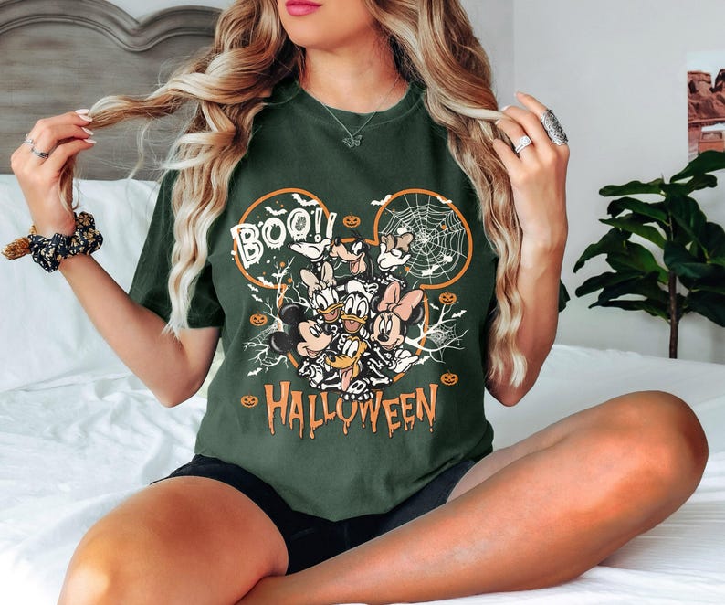 K&ouml;nnte beinhalten: Dunkelgr&uuml;nes T-Shirt mit einem Halloween-Motiv. Das Design zeigt Micky Maus, Minnie Maus, Donald Duck, Daisy Duck und Goofy, mit den Worten "BOO!!" und "HALLOWEEN" in Orange und Wei&szlig;. Das T-Shirt hat einen Rundhalsausschnitt und kurze &Auml;rmel.