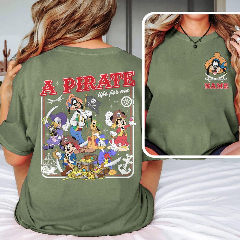 May include: Olive green t-shirt featuring a pirate-themed graphic with various cartoon characters, treasure, and the text "A Pirate life for me." A smaller image of a pirate character is on the front.