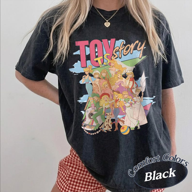 May include: A black acid-wash t-shirt featuring a graphic print with the text "TOY story" in pink and yellow. The design displays a collage of various female characters in colorful outfits, including a camera and a ball, set against a blue background.