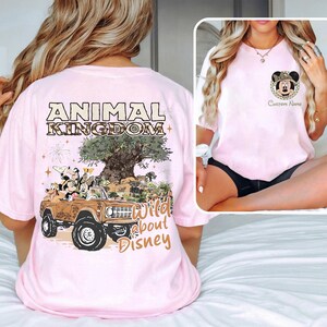May include: A light pink t-shirt featuring a graphic of Mickey Mouse and friends in a safari vehicle with the text "ANIMAL KINGDOM" and "Wild about Disney". The shirt has a crew neck and short sleeves.