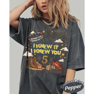 May include: A charcoal gray t-shirt featuring a graphic print with the text I KNEW IT I KNEW YOU, YOU'VE GOT A FRIEND IN ME, and infinity and beyond. The design includes a cowboy boot, a red hat, alphabet blocks, the number 5, and stars.